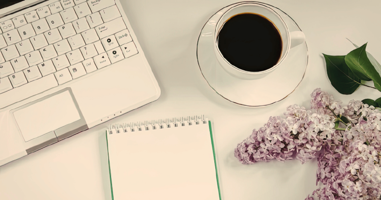 A peaceful desk scene with the corner of a laptop keyboard just visible to the left. An empty notebook and a cup of coffee are centre of the scene and a lilac blossom is on the edge of the right.