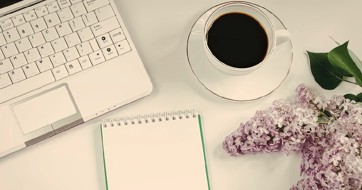 A peaceful desk scene with the corner of a laptop keyboard just visible to the left. An empty notebook and a cup of coffee are centre of the scene and a lilac blossom is on the edge of the right.